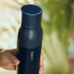 A hand holds the LARQ Bottle in Monaco Blue featuring a blue light at the rim of the bottle cap, palm leaves in the background.