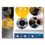A spread from Materialising Colour featuring an image of an overhead view of 6 beakers. 2 of the beakers are filled with a yellow liquid while the remaining four are filled with black liquid. Four of the beakers have been placed on some technical equipment while the last two are placed on a countertop to the left of the viewer.