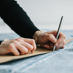 Lifestyle image of a person drawing a line with the Handmade Stainless Steel Pen along a cardboard edge on a piece of denim fabric. Image is cropped to hands.