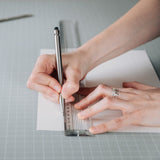 Cropped view of a person’s hands making a mark with a Steel Pencil on a white paper while steadying a clear ruler over the paper which is on a gray cutting mat.