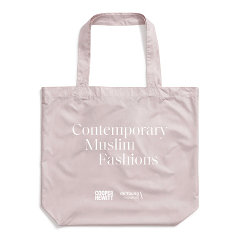 Light purple-gray tote bag made of a slightly shiny material, with white serif text reading "Contemporary Muslim Fashions." Cooper Hewitt logo next to de Young musum logo at bottom center.