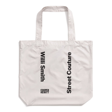 Light gray tote bag made of a slightly shiny material, with vertical bold, black text reading "Willi Smith" facing opposite "Street Couture". Cooper Hewitt logo at bottom left.