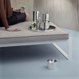 Collection of steel barware on a steel tray, sits on a taupe cushioned bench with a seated person, cut off at the edge of the image, to the left. Another steel vessel sits on the powder blue floor and the wall behind features a pale woodgrain pattern.