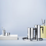 Against a powder blue background, a steel creamer and sugar bowl sit on a white block, and to the right a steel french press and jug sit on a steel tray. A yellow block holding a spouted vessel is cut off the edge of the image.