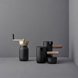 Four sleek black coffee-service vessels on a gray, cornered background; a cylindrical coffee grinder with brass grinding bowl and crank handle, a tall cylindrical lidded milk jug with spout and rubber wood side handle, a flat-topped cylindrical sugar bowl and a tall straight-sided espresso maker with a flat lid, pouring spout and rubber wood side handle