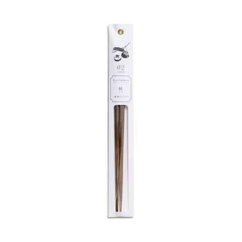A slender, vertical display package of white paper has a dark gray illustration, product information and gold grommet for hanging  above a clear, narrow window containing a pair of partially visible, tapered, persimmon-wood chopsticks.
