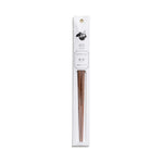 A slender, vertical display package of white paper has a dark gray illustration, product information and gold grommet for hanging  above a clear, narrow window containing a pair of partially visible, tapered, mandarin-wood chopsticks.