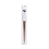 A slender, vertical display package of white paper has a dark gray illustration, product information and gold grommet for hanging  above a clear, narrow window containing a pair of partially visible, tapered, mandarin-wood chopsticks.