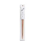 A slender, vertical display package of white paper has a dark gray illustration, product information and gold grommet for hanging  above a clear, narrow window containing a pair of partially visible, tapered, light, natural-wood chopsticks.
