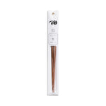 A slender, vertical display package of white paper has a dark gray illustration, product information and gold grommet for hanging  above a clear, narrow window containing a pair of partially visible, tapered, peach-wood chopsticks.