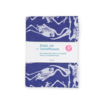 Indigo blue textile with an off white pattern of animal skeletons, folded into a rectangle, wrapped with a white belly band that has teal text and a pink circle logo.
