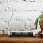 An interior space with a white brick wall and natural wood table surface has a small cylindrical glass tube with silver end caps featuring a small black digital clock set inside the tube, next to a partial view of a clay-potted cactus.