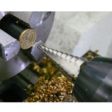 A coin being held in a vice while a drill bit shaves it down. Gold shavings form a pile underneath.