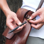 Lifestyle image of hands holding the Black Brassing Portable Ball Point Pen, detaching it from the shackle which is attached to a loop on a leather messenger bag.