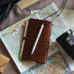 Lifestyle image of the white Brassing Portable Ball Point Pen, lying on a leather notebook on top of a map, surrounded by a camera lens and other papers.